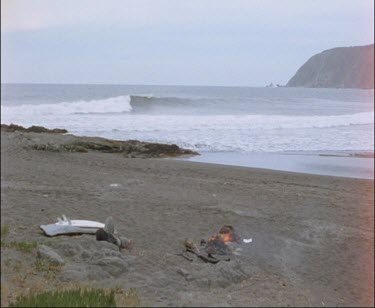 Lone Surfer Guy Lying Down On Beach With Bonfire, Second Man Walks Into Shot