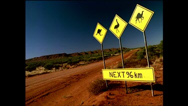 Road signs next to dirt track "Kangaroos, Emus, Camels, next 96 km". Four wheel drive drives by.