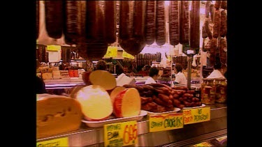 Track past cheese and meat in a market deli, salesman smiles.