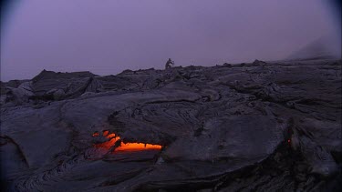 Across lava crust. In the distance a person walks on the volcano. Pan to a vent where lava is erupting. A lot of steam.