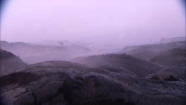 Steam and smoke blowing across screen. Dark volcanic rocky landscape. Eerie. People walking in distance.